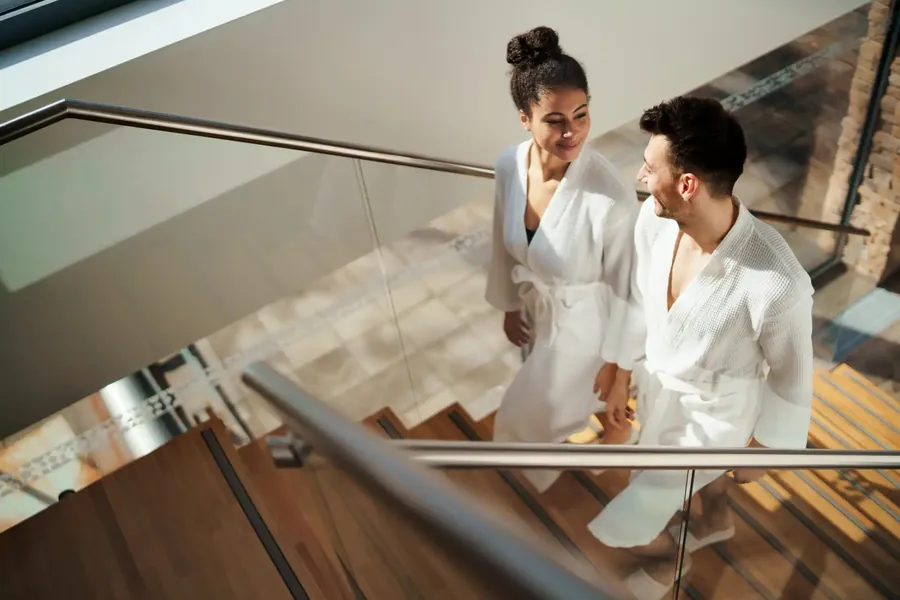 Two guests in white bathrobes walking up a wooden staircase in a spa
